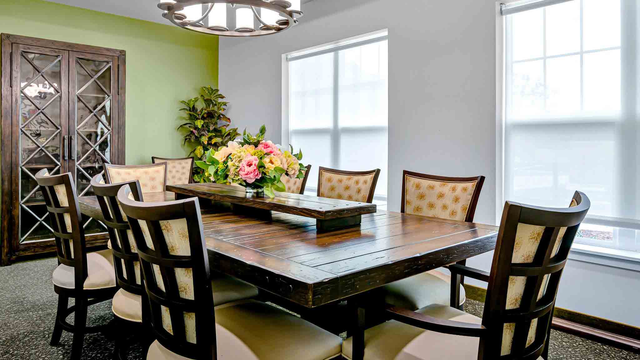 A dining room with a dark wooden table, eight cushioned chairs, a flower arrangement centerpiece, a chandelier, a green accent wall, a large cabinet, and two windows with white blinds.