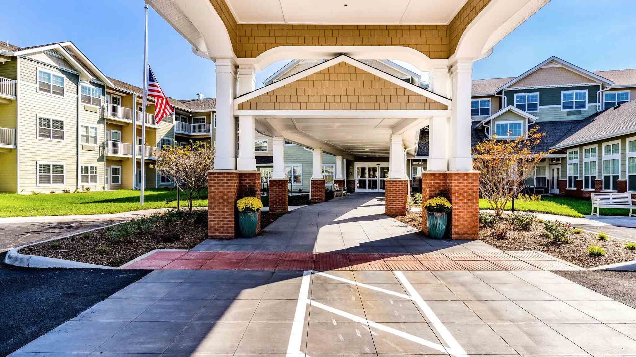 Wide exterior view of a senior living facility entrance with a covered driveway, brick columns, potted plants, American flag, and landscaped grounds on a sunny day.