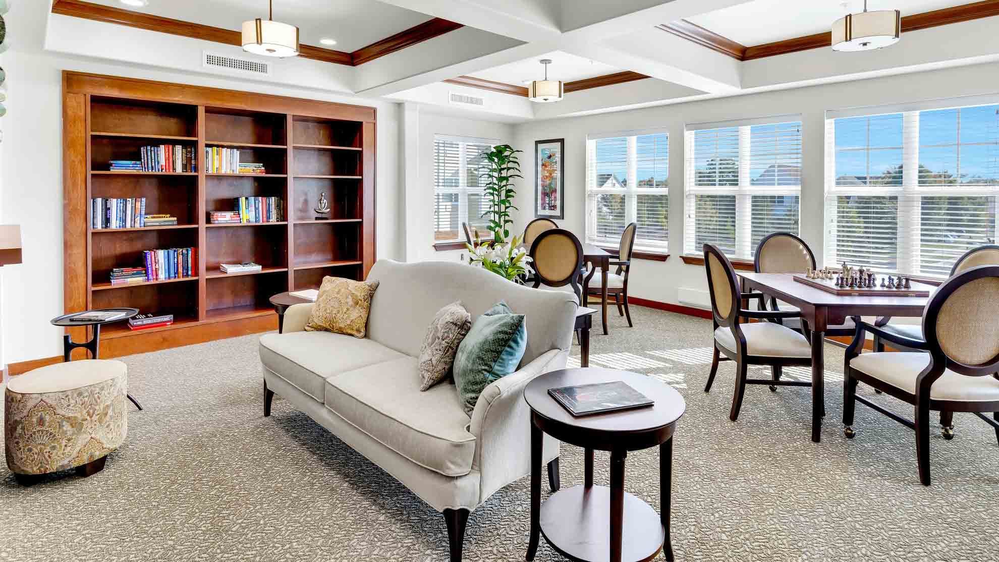 A bright, spacious lounge with a white sofa, side tables, round dining tables with chairs, a large bookshelf with books, and large windows allowing natural light to fill the room.