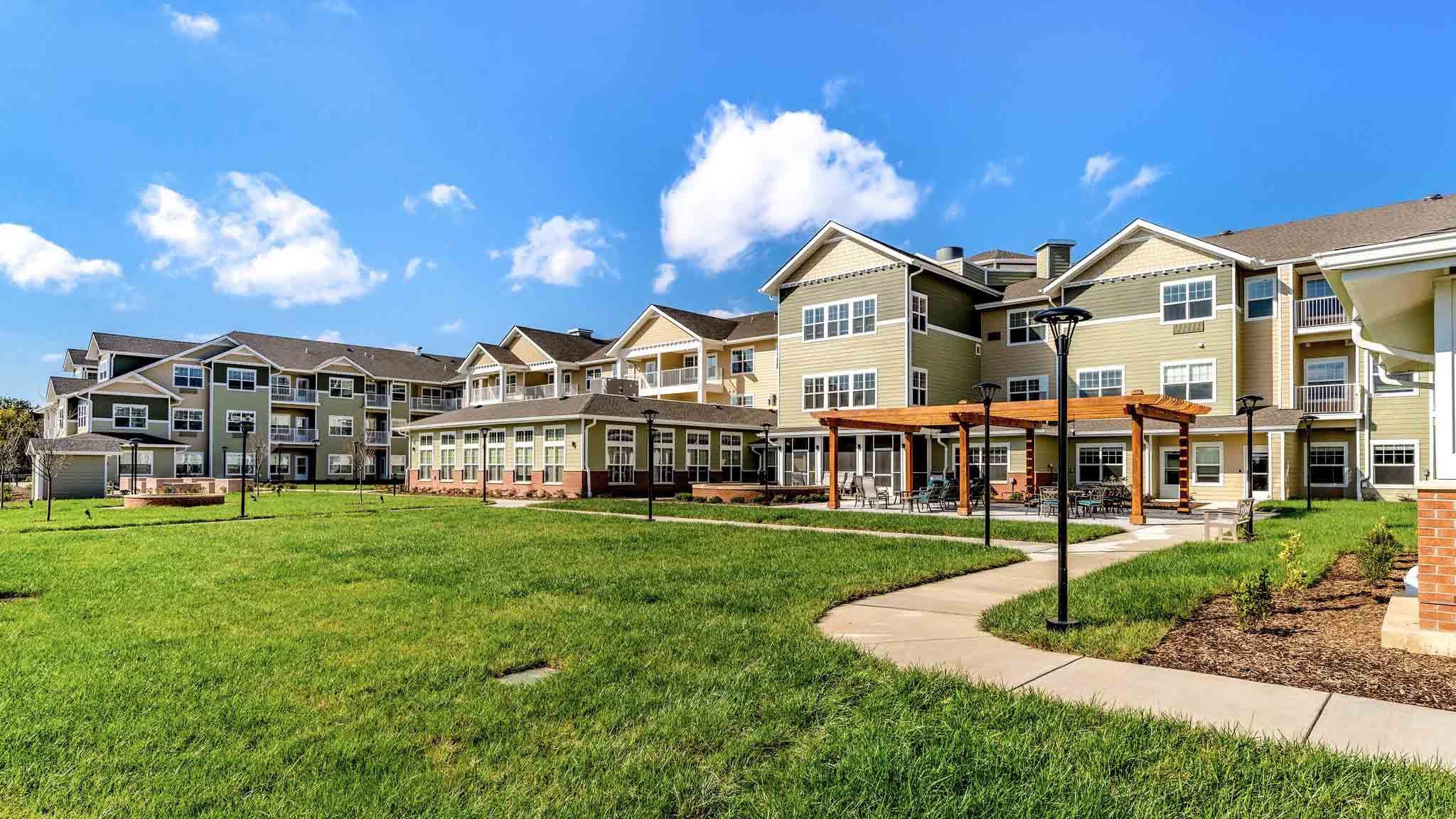 A modern apartment complex with multiple stories, beige and green siding, large windows, balconies, and a covered patio area surrounded by green lawns and a sidewalk under a bright blue sky with scattered clouds.
