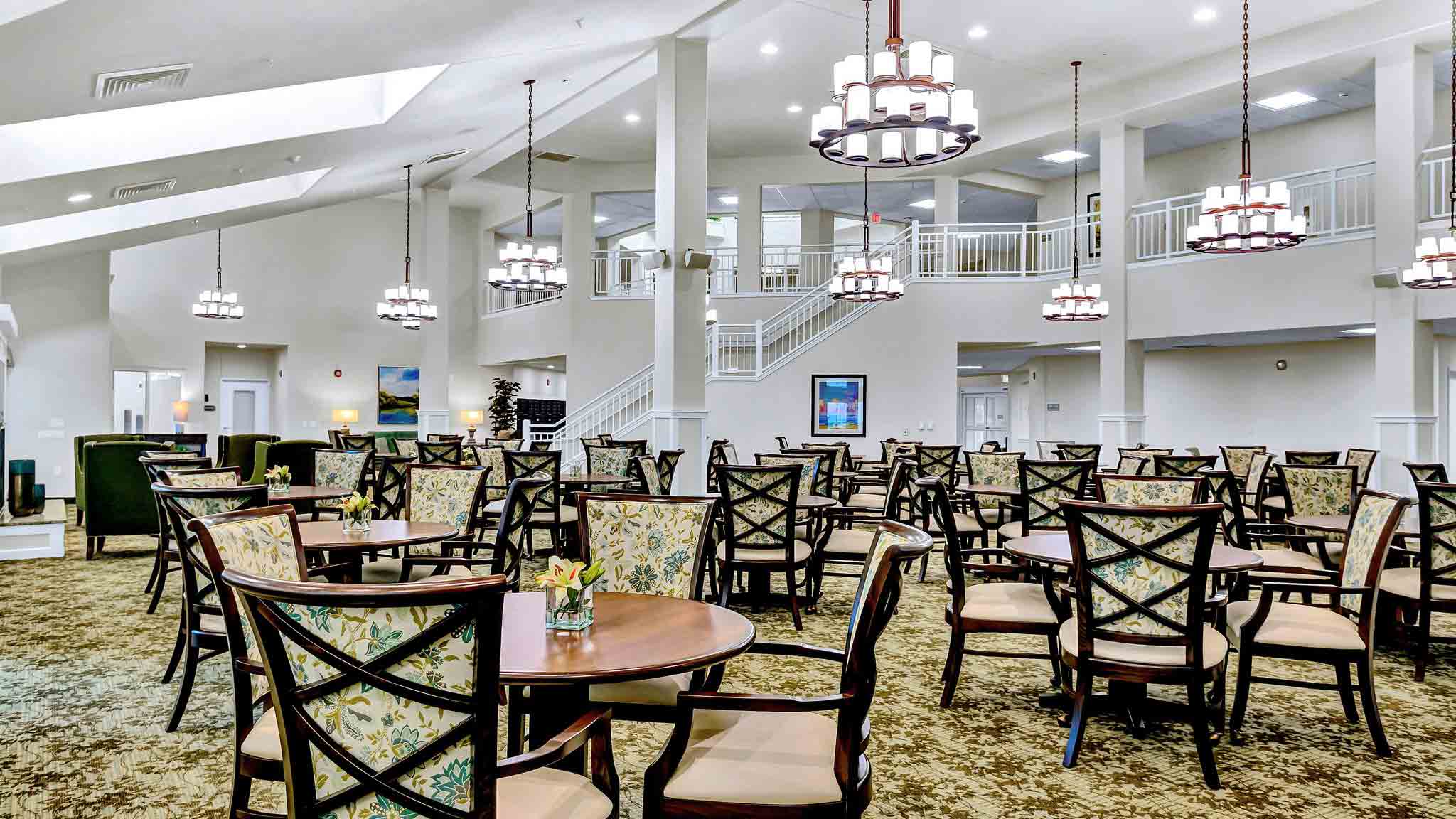 Spacious, well-lit dining area with multiple round tables and cushioned chairs. Elegant chandeliers hang from the ceiling, and a staircase with a white railing leads to an upper level. Large windows let in natural light.