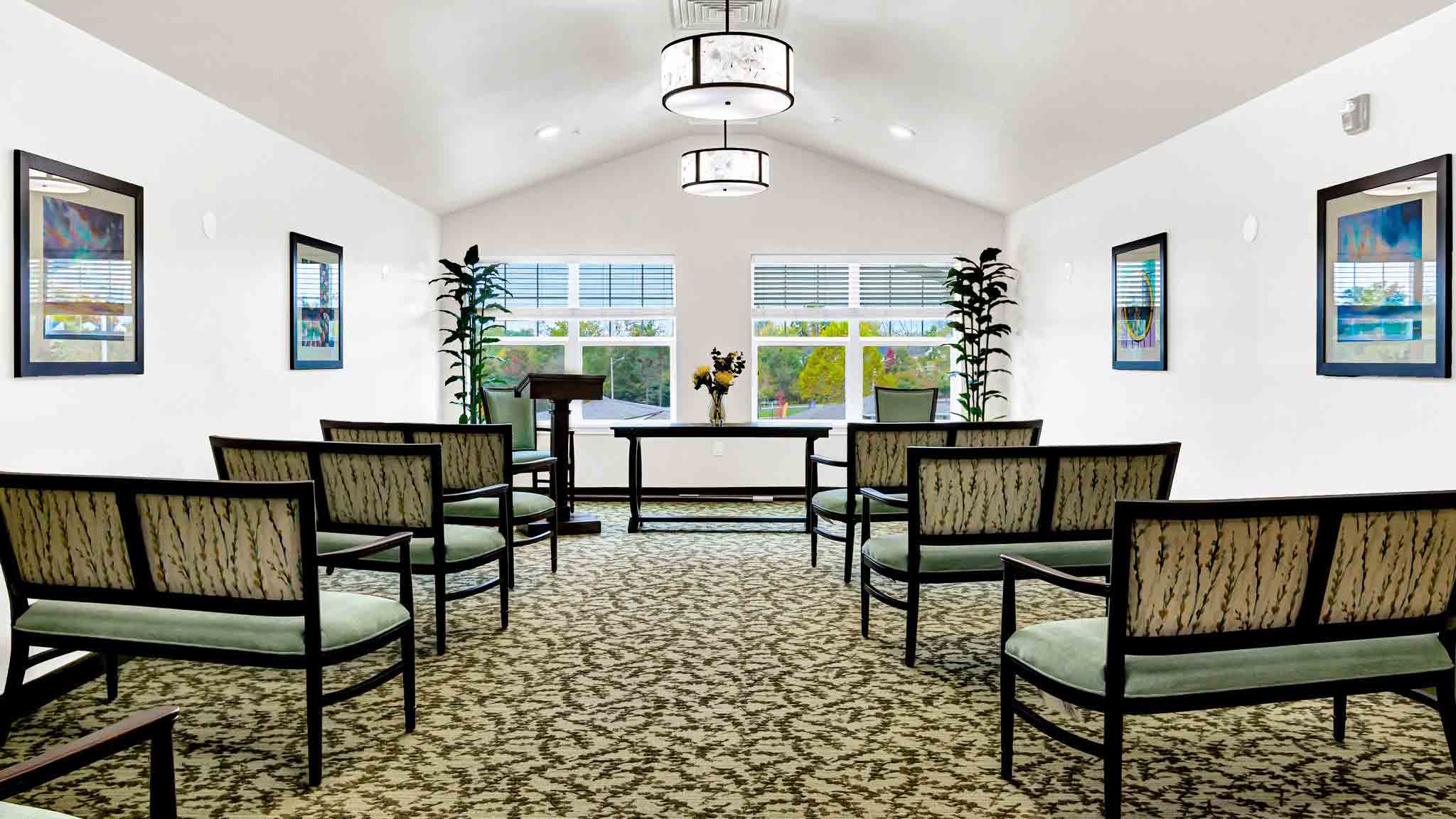 Small, bright chapel with patterned carpet, rows of cushioned chairs, a lectern, and a table with flowers. Large windows at the far end let in natural light; framed art and plants decorate the walls.