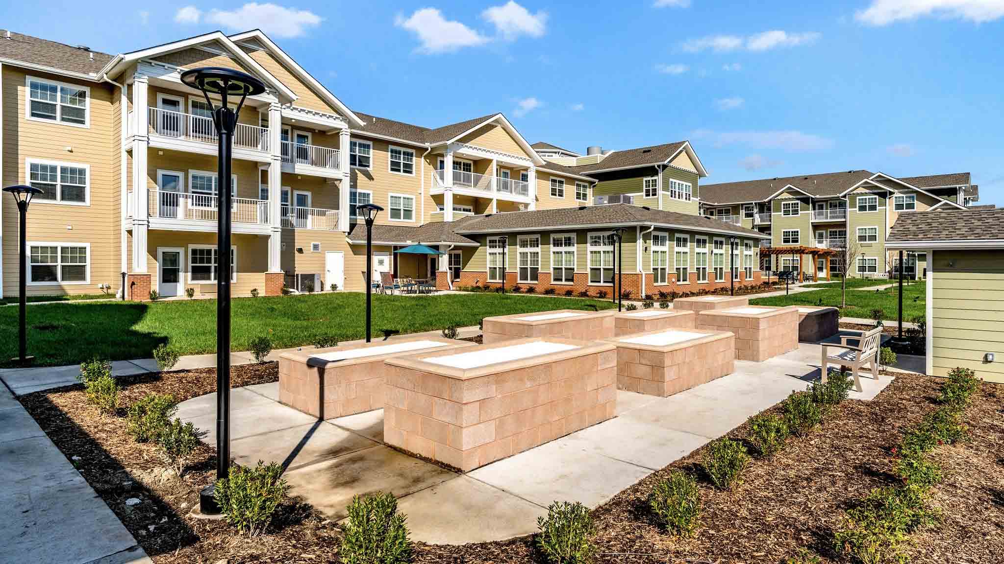 A modern apartment complex with light yellow siding, multiple balconies, manicured lawns, outdoor seating areas, and raised brick planter beds under a blue sky with scattered clouds.