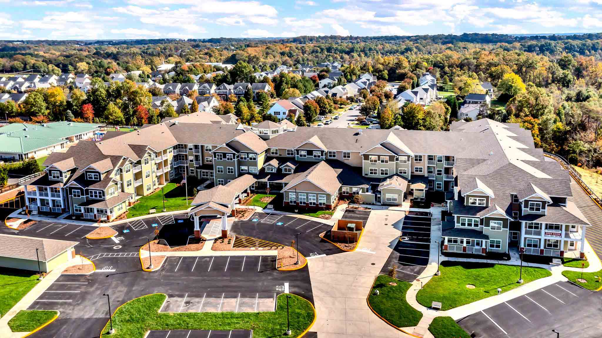 Aerial view of a large apartment complex with multiple connected buildings, surrounded by parking lots, trees, and residential homes in the background on a sunny day.
