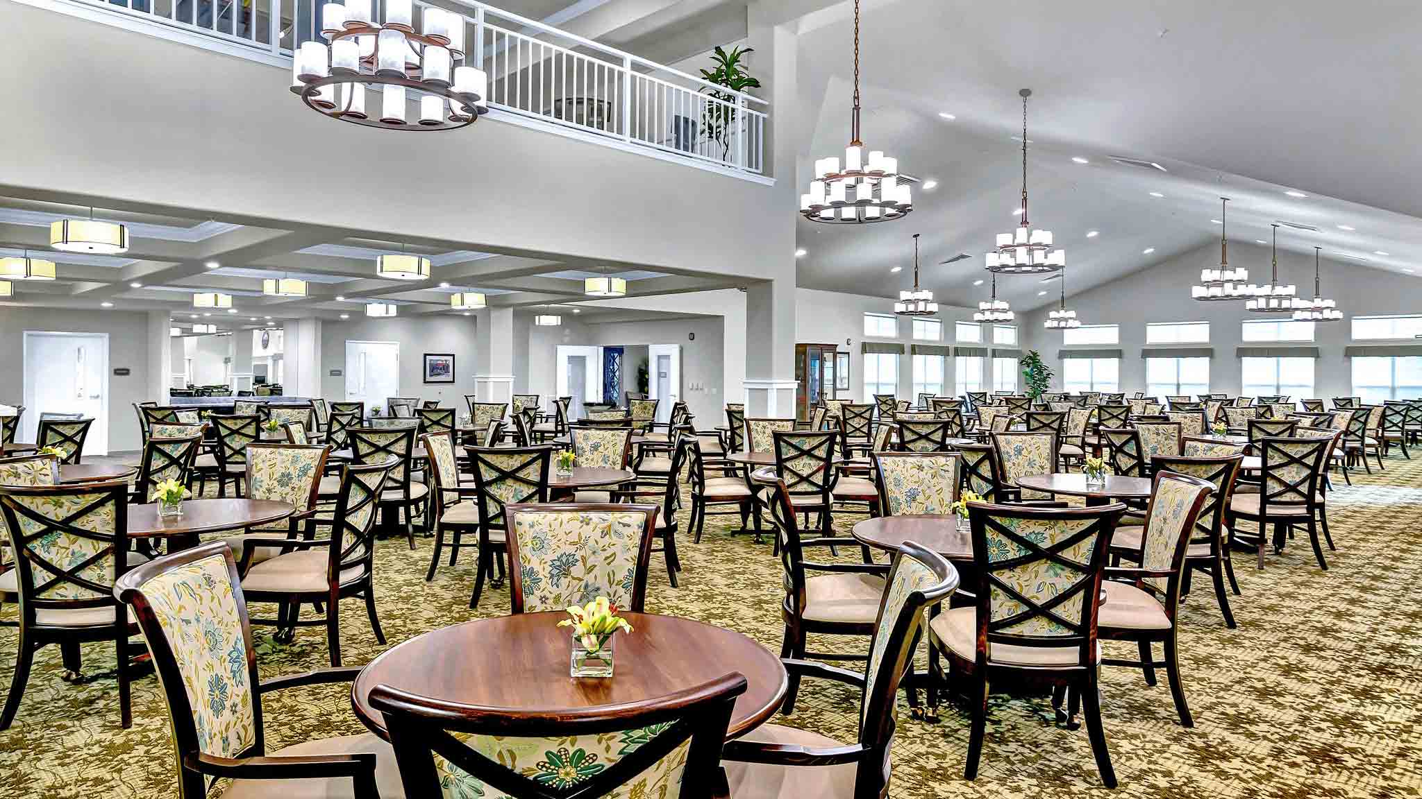 A spacious, well-lit dining area with many round wooden tables and patterned chairs. Modern chandeliers hang from the ceiling, and a small vase with yellow flowers sits on a table in the foreground. Large windows let in natural light.