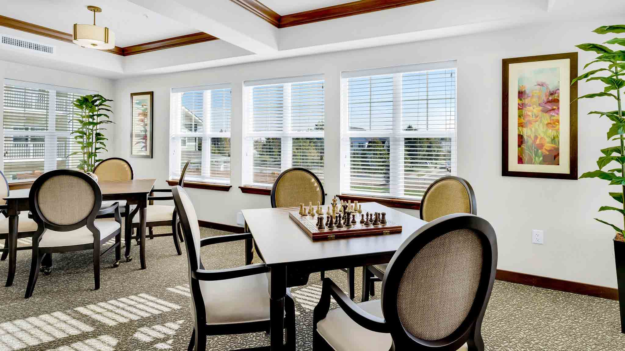 Bright room with large windows, several tables and chairs, a chessboard set up on one table, green plants, framed artwork on the wall, and natural sunlight streaming in.