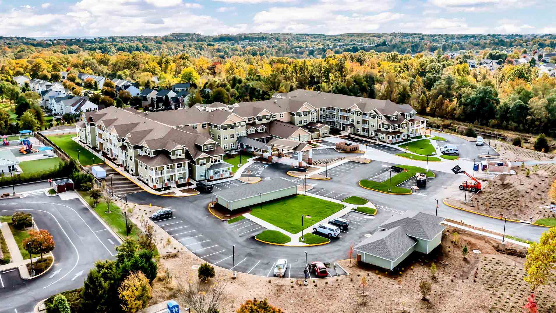 Aerial view of a large, modern residential or assisted living complex surrounded by trees with autumn foliage, parking lots, landscaped areas, and nearby suburban houses under a partly cloudy sky.