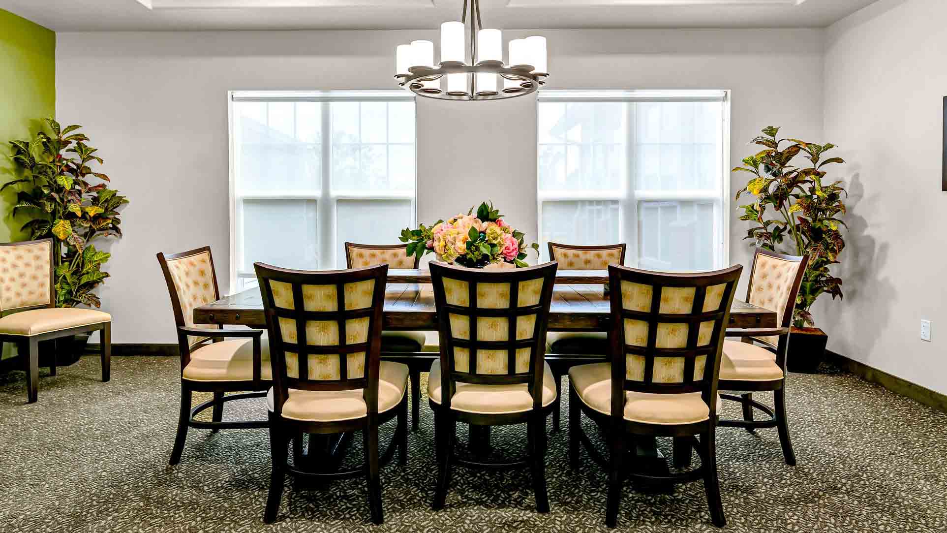 A dining room with eight cushioned chairs around a wooden table, a floral centerpiece, two potted plants near large windows, and a modern chandelier hanging above. The room has neutral walls and patterned carpet.