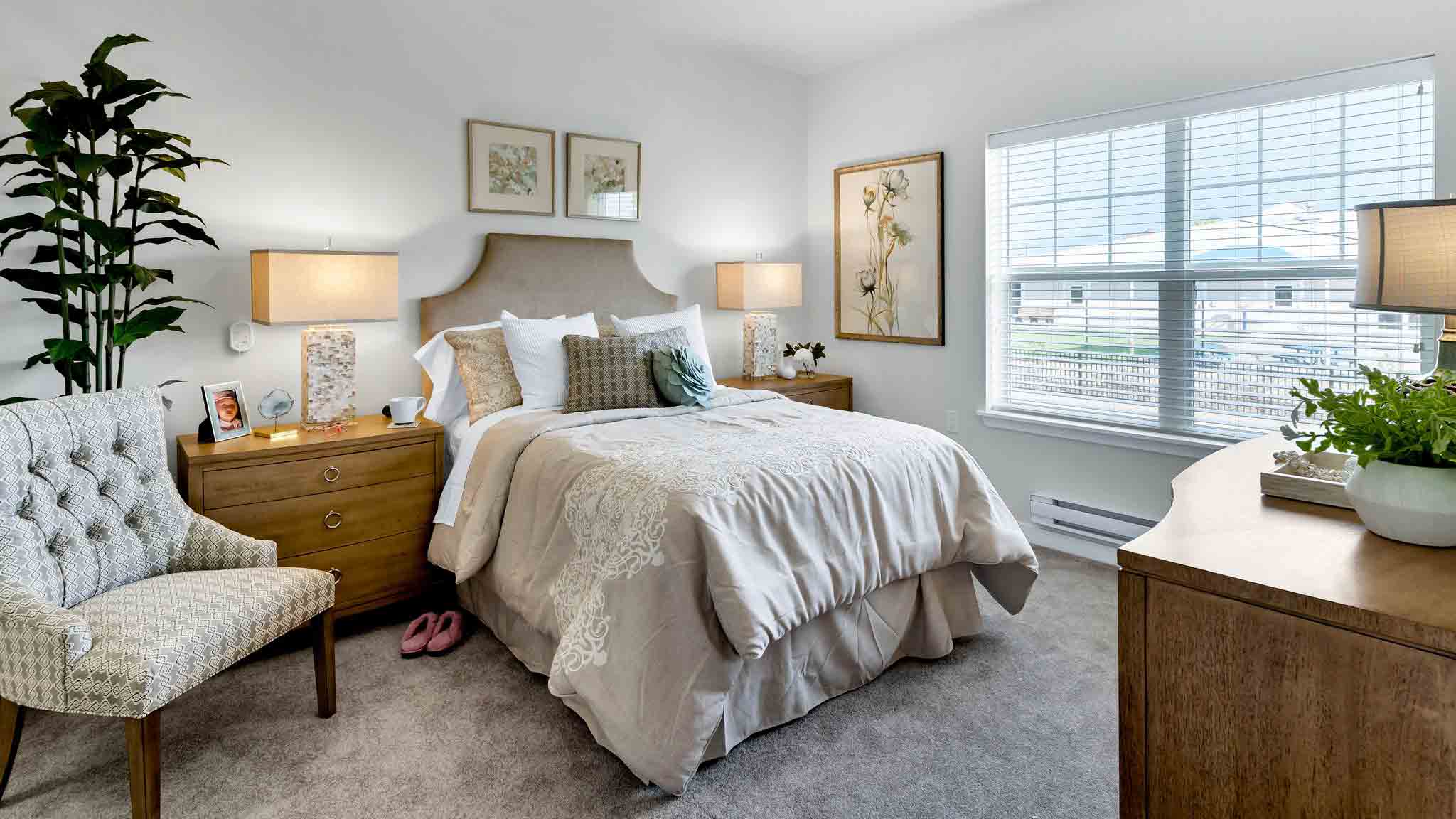 A cozy, well-lit bedroom with a neatly made bed, two wooden nightstands with lamps, a cushioned chair, artwork on the walls, a large window with blinds, and a tall plant in the corner.