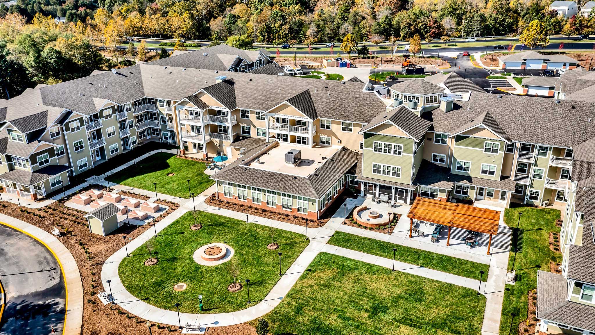 Aerial view of a large, multi-story residential building with landscaped courtyards, walking paths, fire pits, and outdoor seating areas surrounded by green lawns and trees.