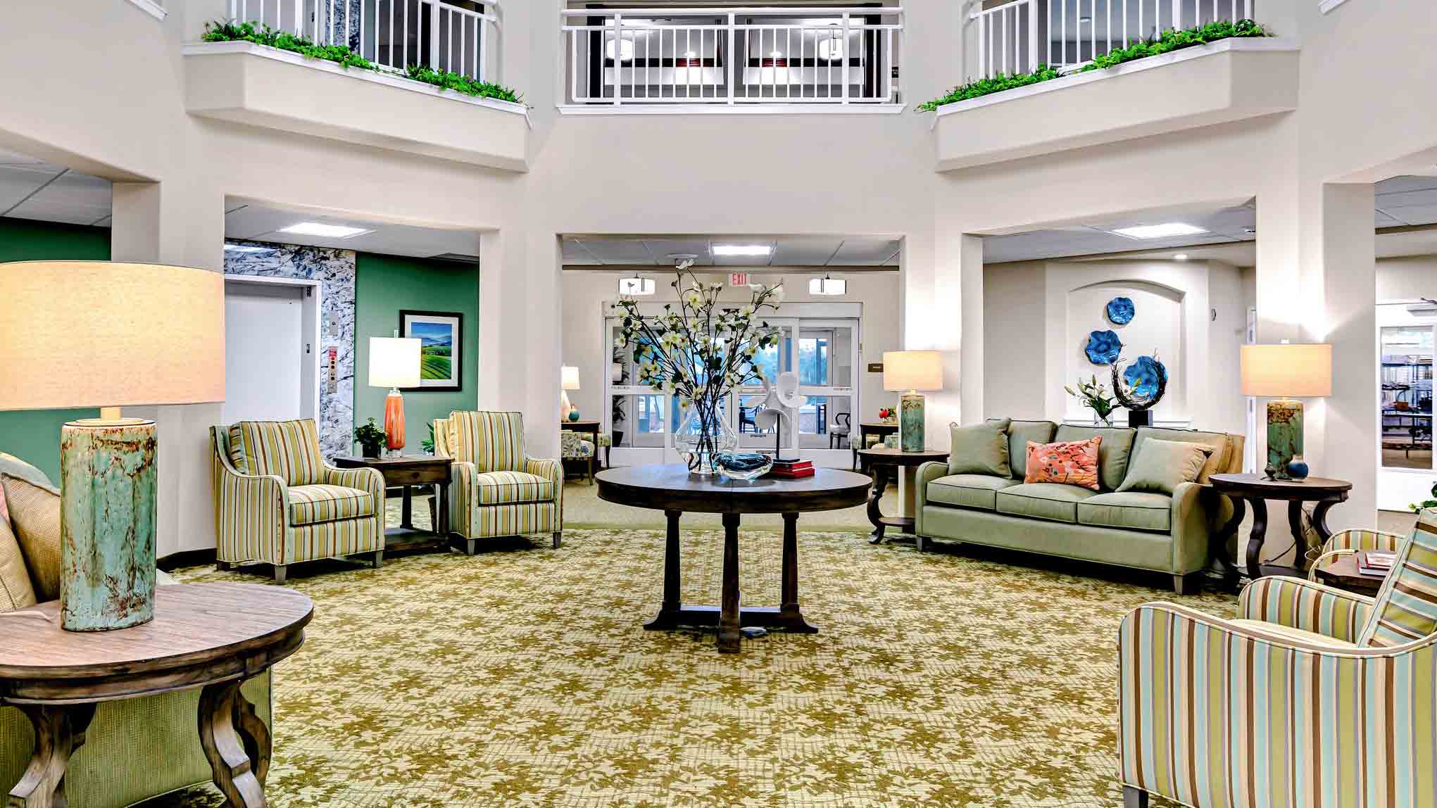 A spacious, brightly lit lobby with green patterned carpet, sofas, armchairs, lamps, and a round table with a floral arrangement in the center. The area features high ceilings and decorative plants.