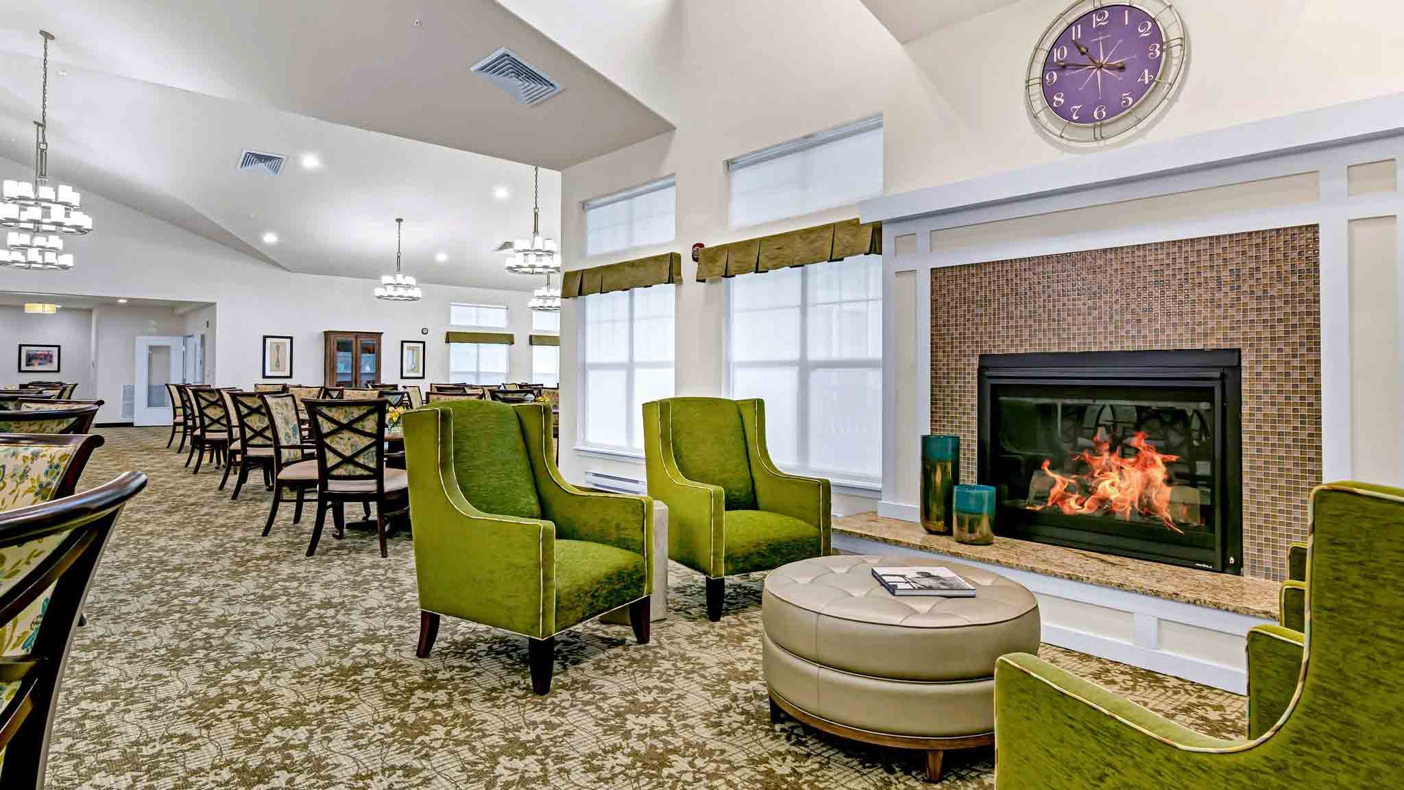 A spacious, bright lounge area with green armchairs around a round ottoman, a lit fireplace, and a large wall clock above. In the background, there are dining tables and chairs under modern chandeliers.