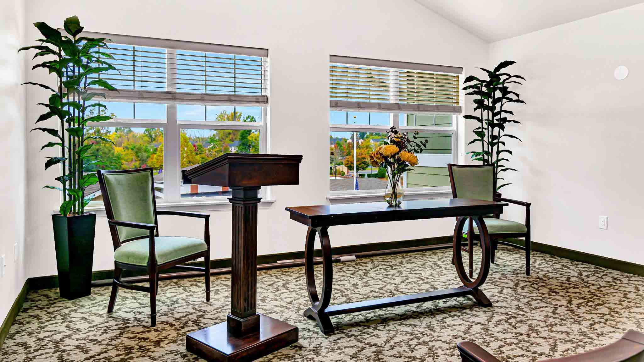 A bright room with two green chairs, a dark wood podium, a matching table with a flower arrangement, large windows, and two tall potted plants on each side. The floor is carpeted with a floral pattern.
