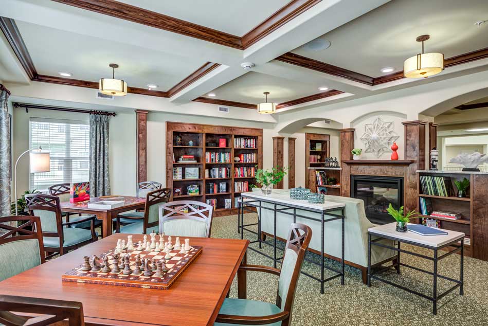 A cozy, well-lit library and game room featuring a chessboard on a wooden table, cushioned seating, bookshelves filled with books, and a fireplace. The space has wooden beams on the ceiling and is decorated with plants and a window with curtains.