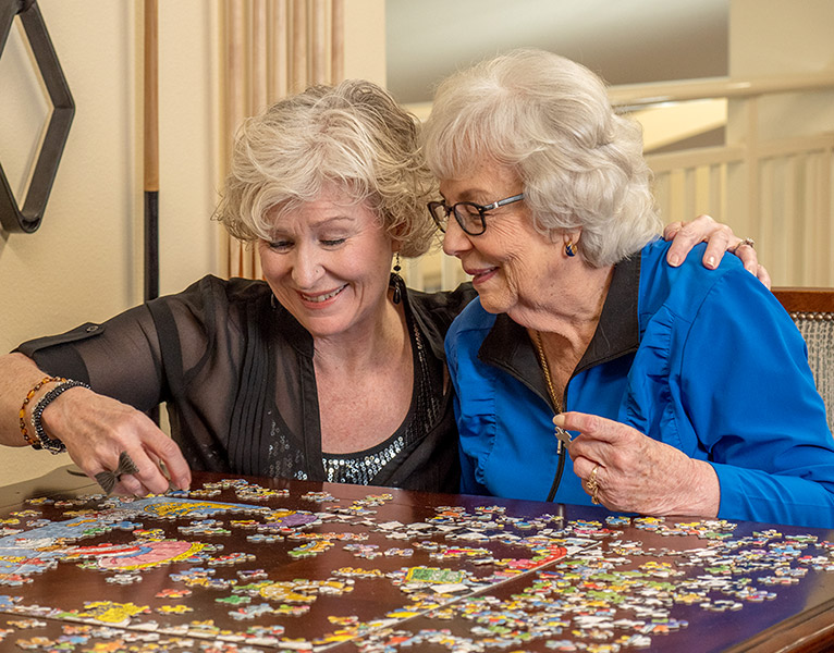 Two elderly women are seated at a table, smiling and working together on a colorful jigsaw puzzle. One woman is placing a piece while the other looks on encouragingly, her arm around her companion. The scene conveys a sense of warmth and companionship.