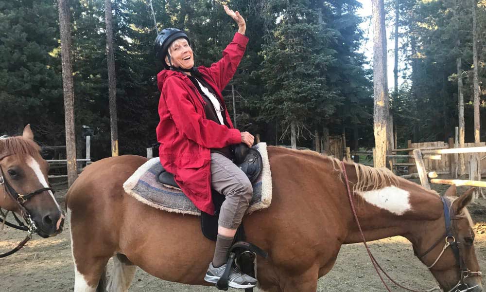 A smiling person in a red jacket and helmet sits on a brown and white horse, waving enthusiastically. Another horse is seen in the background. The scene is set in a forested area with tall trees and a wooden fence.