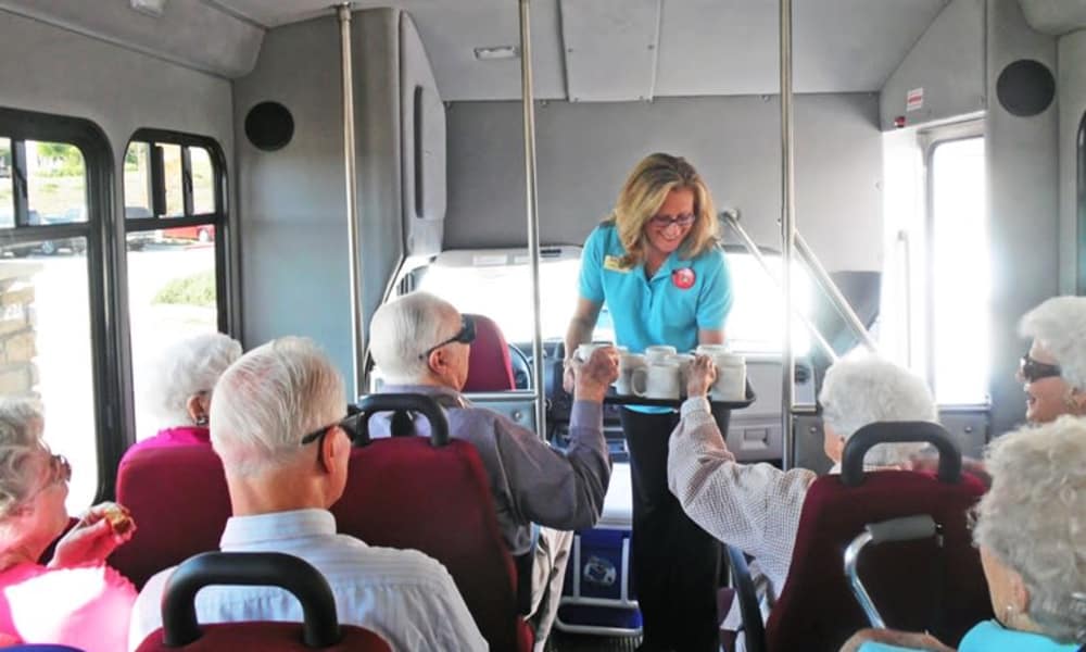 A bus driver with glasses and a blue polo shirt hands a tray with drinks to a group of elderly passengers seated in a bus. The passengers, dressed casually, appear to be enjoying their trip, smiling and chatting with each other.