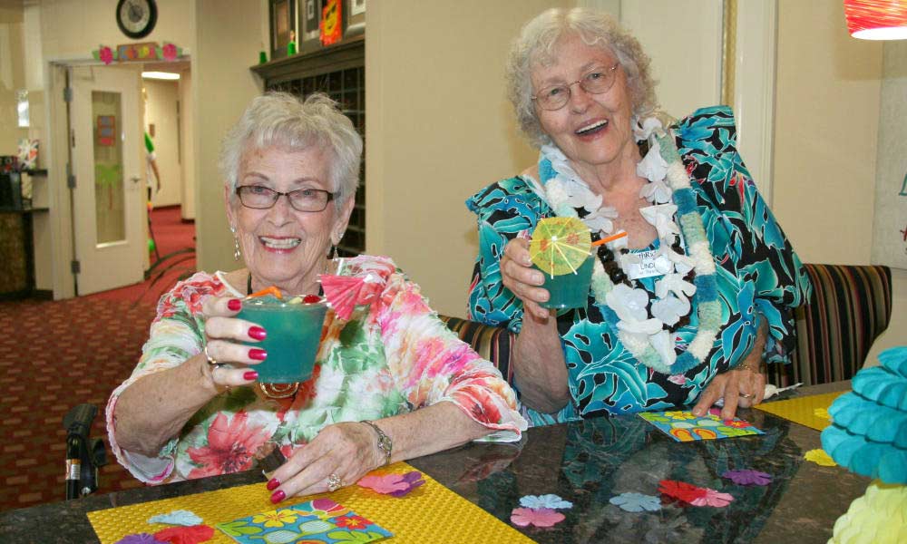 Two elderly women are sitting at a table, smiling and holding up colorful drinks garnished with umbrellas. They are dressed in festive attire, including floral patterns and leis, suggesting a tropical or Hawaiian theme. The background shows a decorated indoor setting.