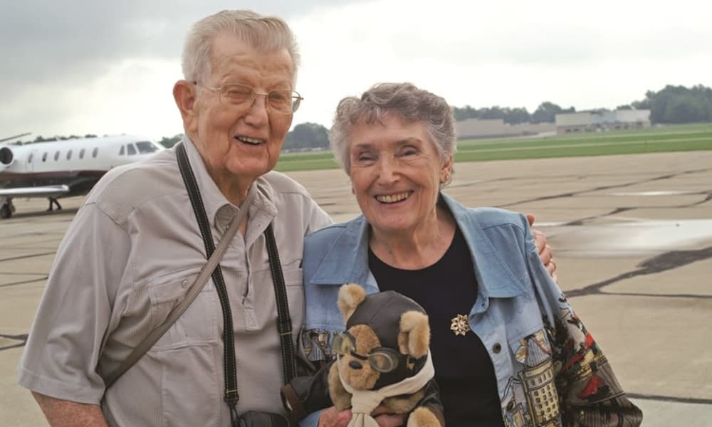 An elderly man and woman smile warmly at the camera while standing on an airstrip with a private jet in the background. The man holds a teddy bear dressed as an aviator. The woman wears a light blue jacket, and both appear happy and cheerful.