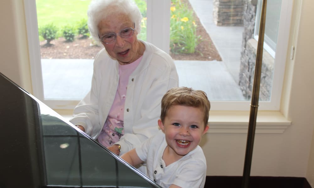 An elderly woman with white hair, wearing glasses and a white cardigan, sits next to a smiling young boy with short blonde hair who is wearing a white shirt. They are both seated at a piano, with a window showing greenery in the background.