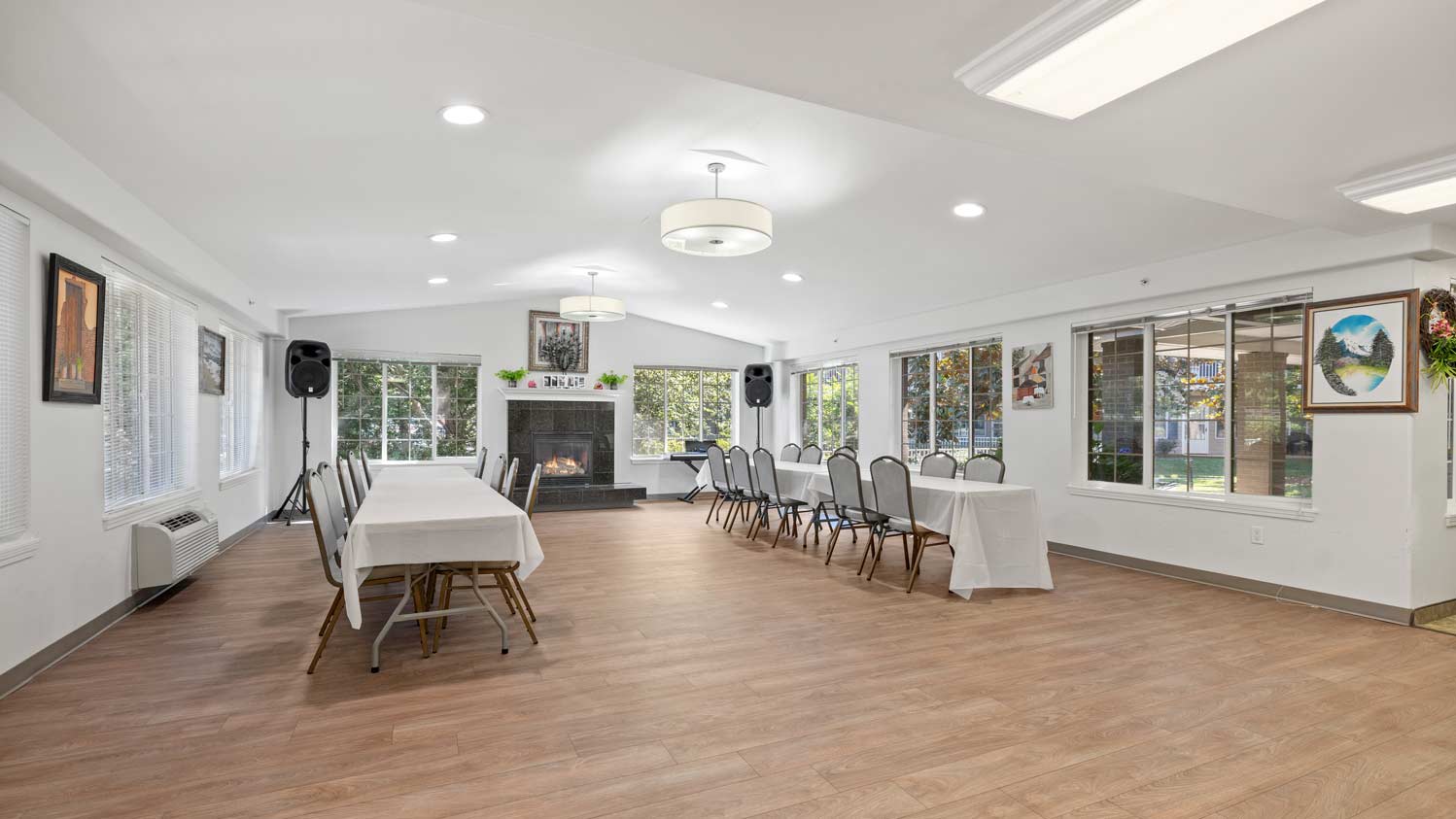 A spacious room with wooden flooring featuring a fireplace in the center of one wall. Several tables are set up with white tablecloths and gray chairs arranged around them. Large windows let in natural light, and various framed pictures adorn the walls.