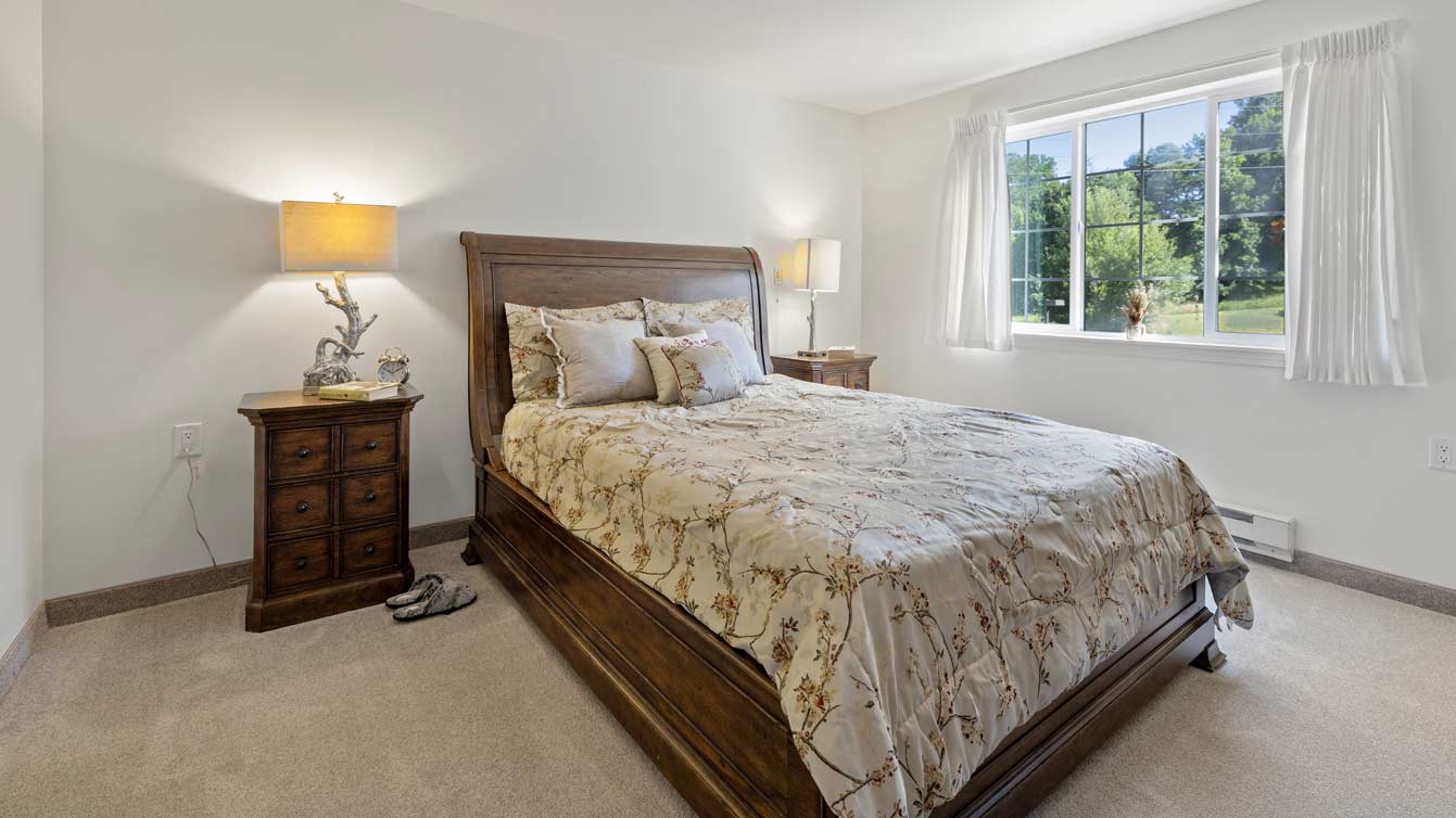 A cozy bedroom features a wooden bed with floral bedding, flanked by two wooden nightstands with lamps. Sunlight spills in from a large window with white curtains, illuminating the room. A pair of slippers rests on the beige carpet near the bed.