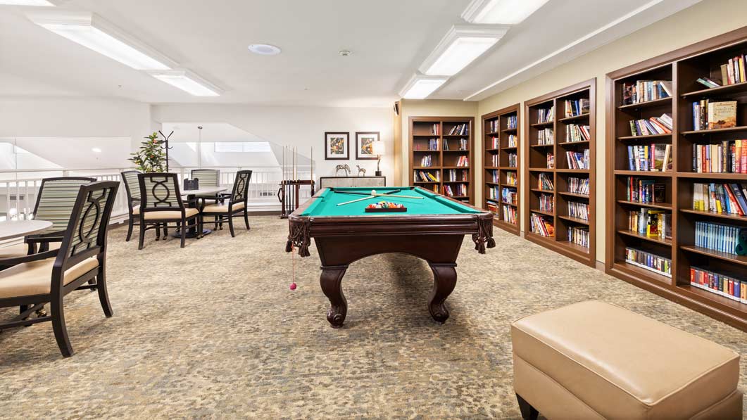 A spacious room with a pool table in the center, surrounded by bookshelves filled with books. There are several chairs and tables arranged around the room. The space is well-lit with modern light fixtures and has a neutral-colored carpet and walls.