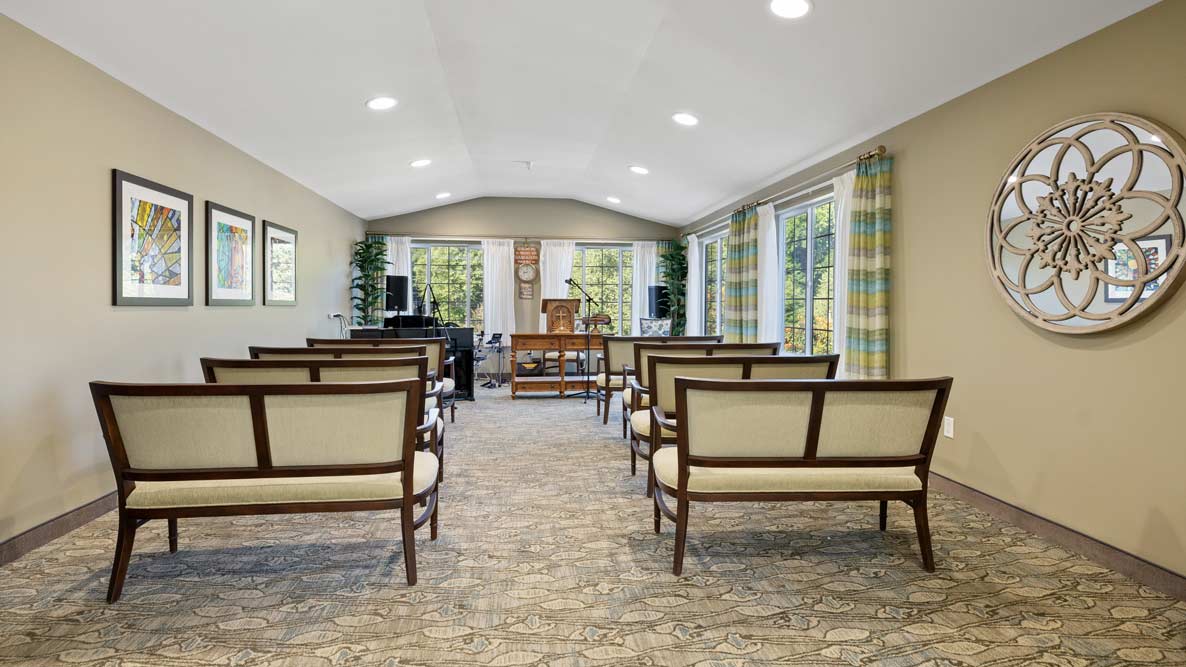 A small chapel or meeting room with rows of wooden benches upholstered in light fabric. The room features large windows with striped curtains, framed artwork on one wall, a decorative circular mirror, and various plants. The space is well-lit with ceiling lights.