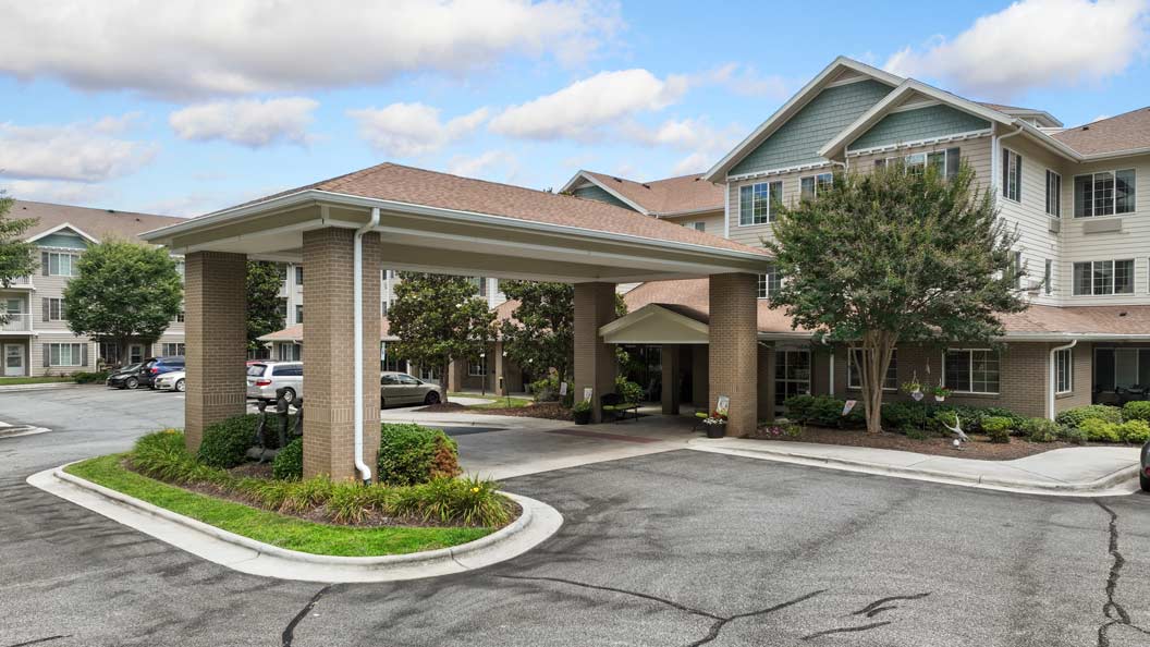 A large residential building with multiple floors featuring light-colored siding and a brown roof. An extended covered entrance supported by brick columns leads to the main door. Several windows and landscaped greenery surround the clean, paved driveway.