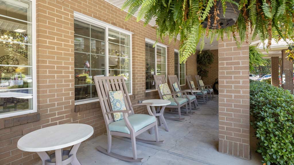 A covered porch with several wooden rocking chairs arranged in a row along a brick wall. Each chair has a cushion and a patterned pillow. Round white tables are placed between some chairs. Green ferns hang from the ceiling, and there are shrubs nearby.