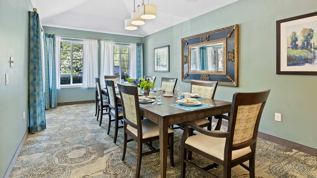 A dining room with a rectangular wooden table set for six. The table is surrounded by upholstered chairs. A large framed mirror hangs on the green wall, and a window with white curtains lets in natural light. A painting and ceiling lights are also visible.