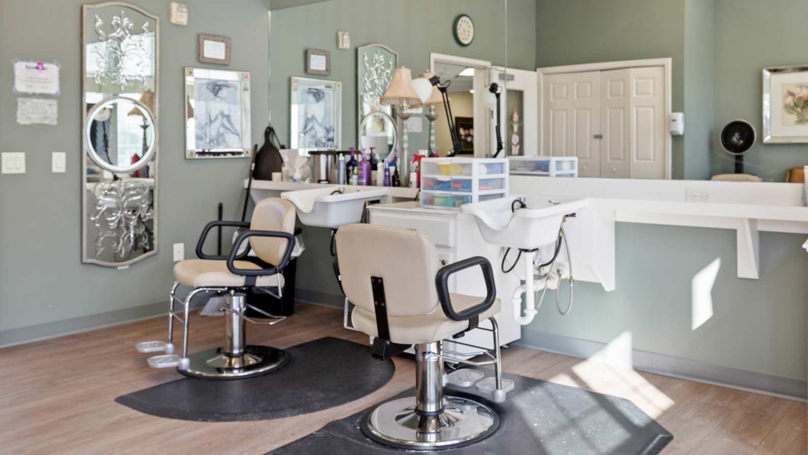 A well-lit hair salon featuring two beige styling chairs in front of mirrors and hair washing sinks. The counters hold various hair products and tools. The walls are a calming green, adorned with decorative mirrors and framed artwork.