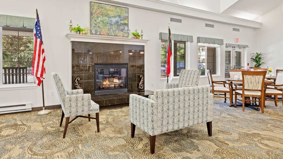 A cozy living room with patterned armchairs facing a fireplace. The room features a large rug, windows with green valances, American flag on left, and a table with chairs on the right. Plants and artwork adorn the mantel and walls, adding to the homey atmosphere.