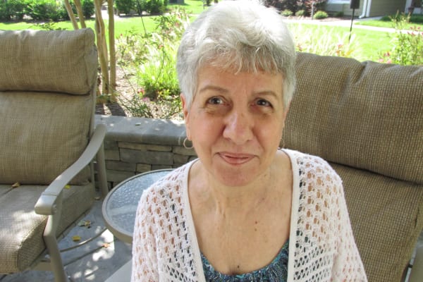 An elderly woman with short gray hair sits outdoors on a cushioned chair, smiling gently. She wears a white crochet cardigan over a patterned blue blouse. The background shows a green lawn and some lush plants, suggesting a garden setting.
