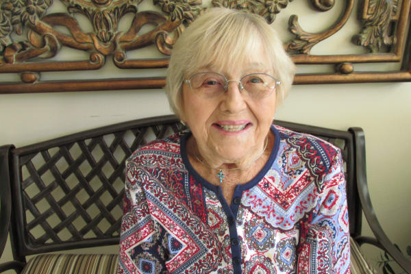 An elderly woman with short white hair and glasses smiles while sitting on a decorative bench. She is wearing a colorful patterned blouse and a small cross necklace. The background features an intricately carved wood and metal wall decoration.