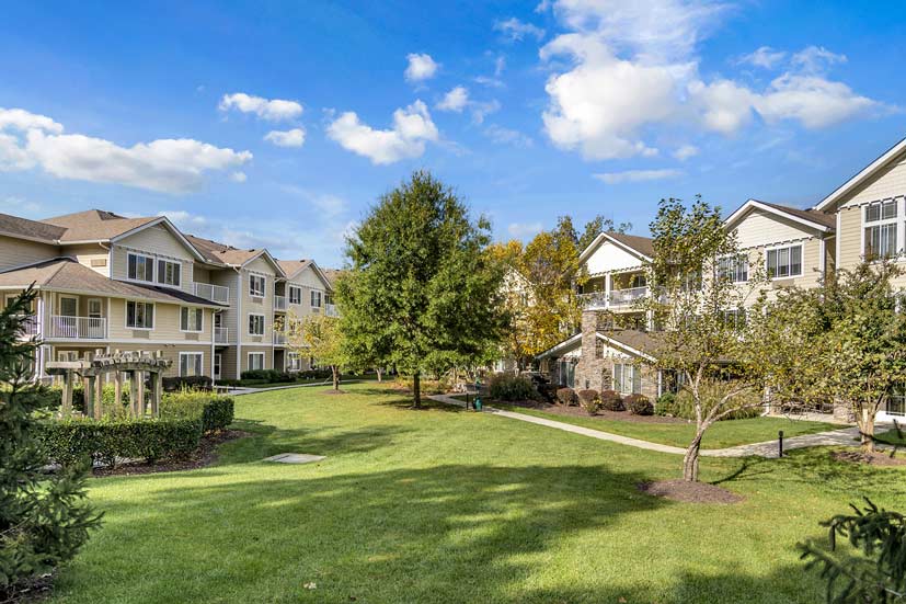 A beautifully landscaped courtyard flanked by modern, multi-story senior living apartment buildings. The area features well-kept lawns, trees, and walking paths under a clear blue sky with a few clouds. The buildings have light-colored exteriors and numerous windows.