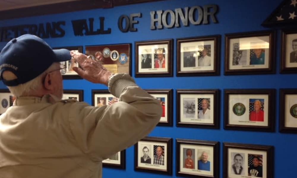 An elderly man in a beige jacket and blue cap salutes a 