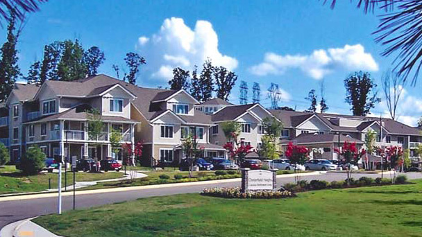 A view of a suburban neighborhood with modern, multi-story houses dedicated to senior living. The residences are situated on a curved street, surrounded by manicured lawns and trees. Flowering bushes line the roadside. A signboard on the lawn is visible under a blue sky with scattered clouds.