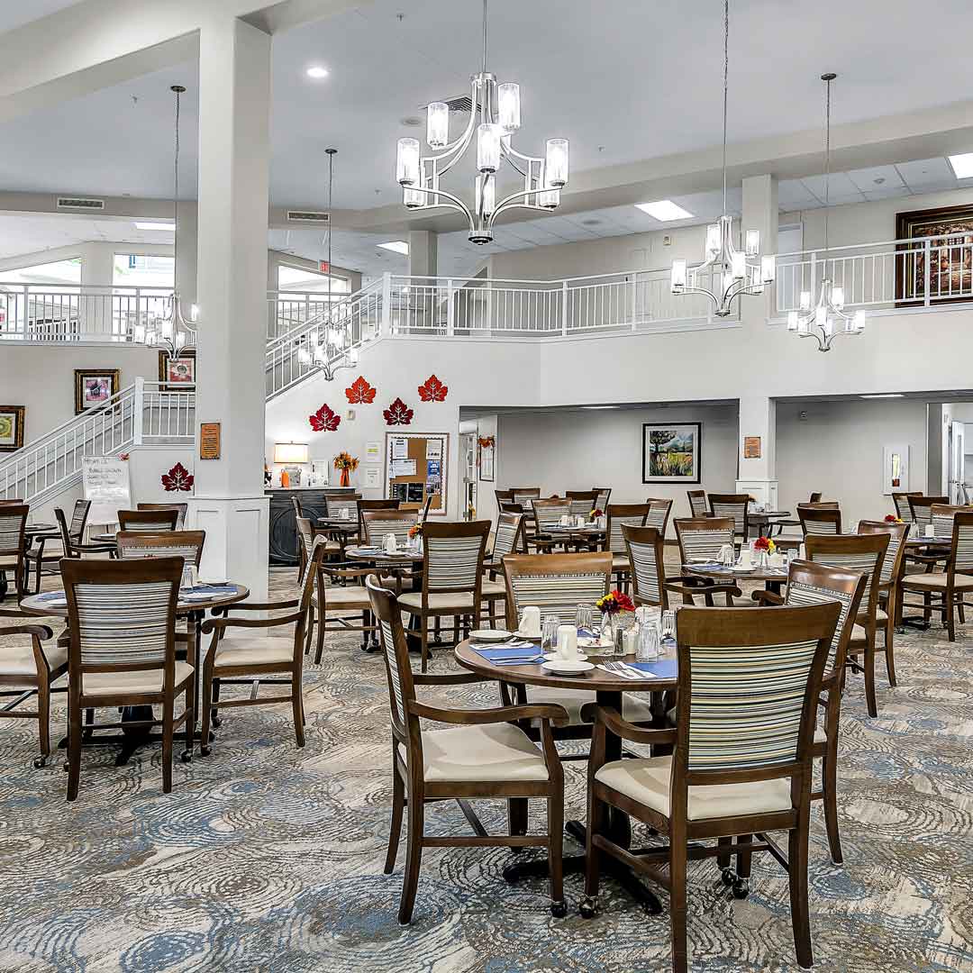 A spacious dining room with several round tables and wooden chairs, each set with plates and cutlery. The area is well-lit by modern chandeliers and has a neutral palette. Walls are adorned with autumn leaf decorations and framed artwork.