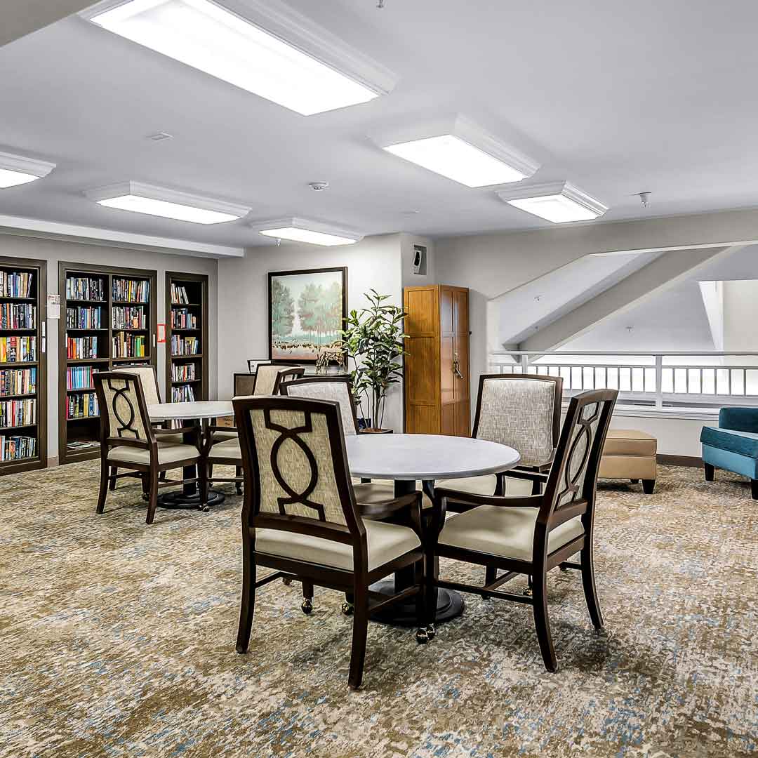 A well-lit library room with bookshelves and a round table surrounded by four upholstered chairs. There's art on the wall, a potted plant nearby, and additional seating, including a teal armchair, on a patterned carpet.