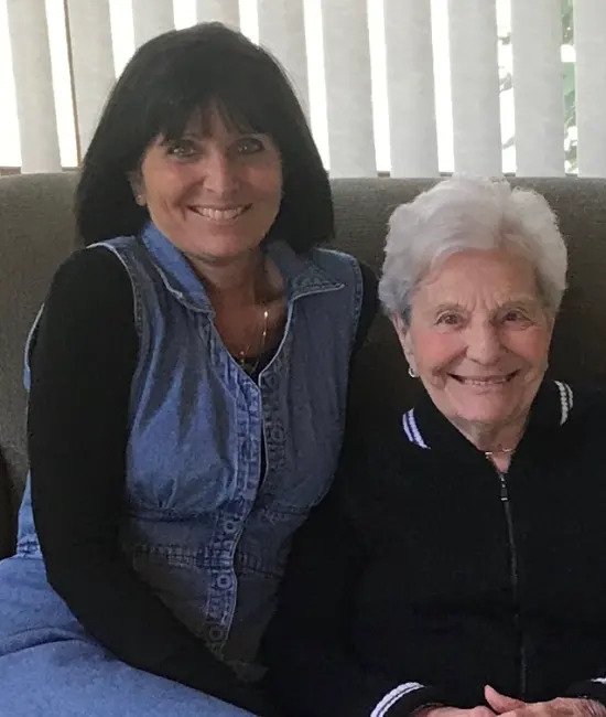 Two women are sitting on a couch, smiling at the camera. The woman on the left has dark hair and is wearing a denim vest over a black long-sleeve shirt. The woman on the right has white hair and is wearing a black jacket with a white collar. Vertical blinds are in the background.