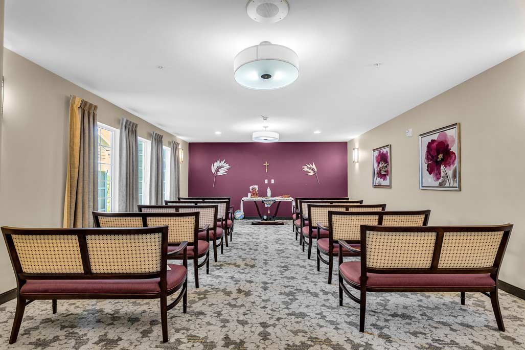 A small chapel with rows of wooden chairs having woven backs and red cushions, facing an altar. The altar is adorned with a cloth and has a cross on the wall behind it. The room features beige walls with burgundy accents and artwork with floral themes, ideal for nearby senior apartments for rent.