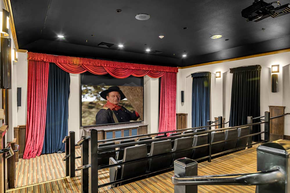 A small, private movie theater with a projector screen showing an image of a man in a hat and coat. The theater has several rows of black cushioned seats, wooden paneling, and is decorated with red curtains around the screen. The lighting is dimmed.