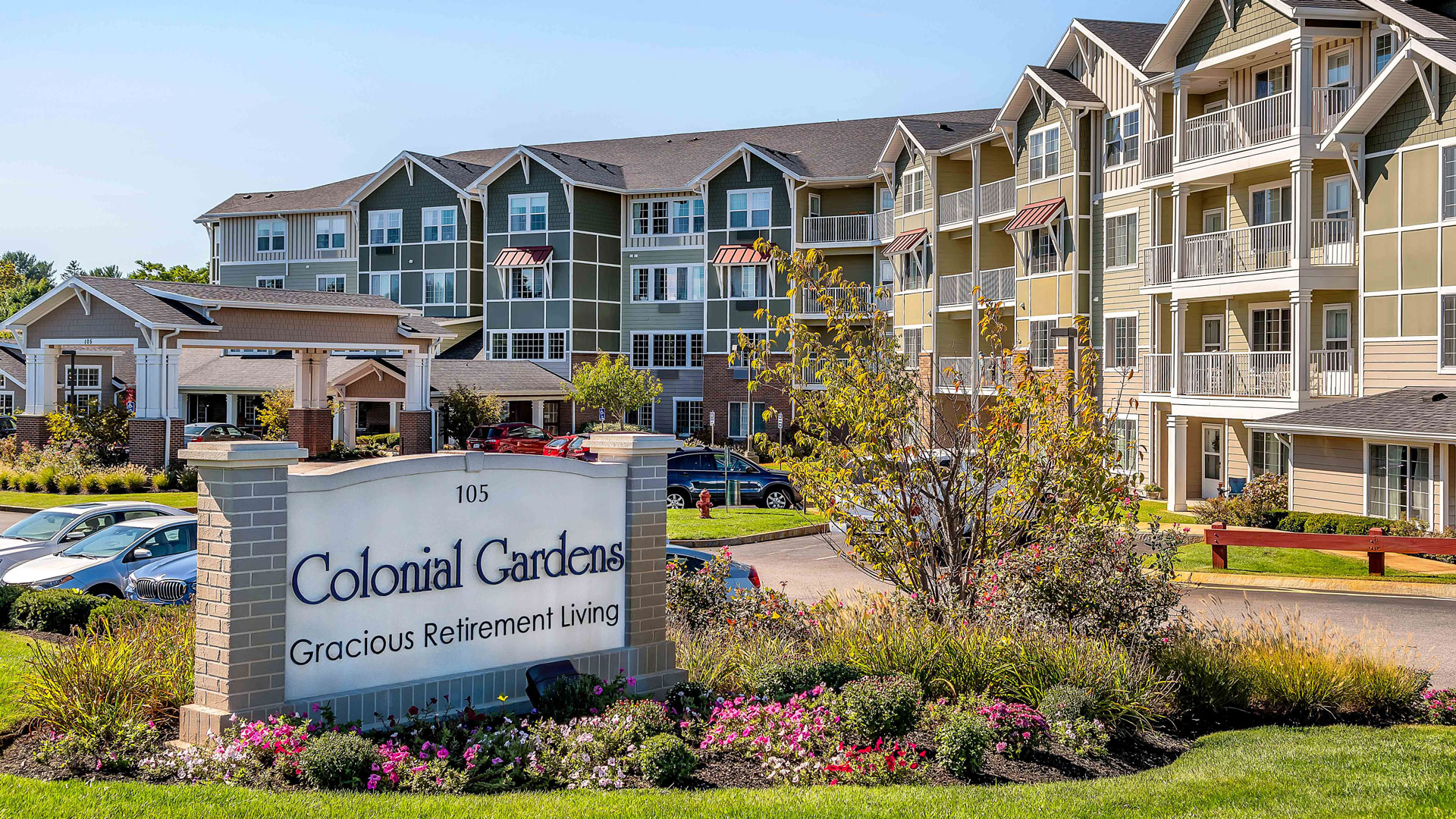 A large, multi-story retirement living complex with balconies and well-maintained landscaping; a sign in front reads “Colonial Gardens Gracious Retirement Living.” Several cars are parked in the lot.