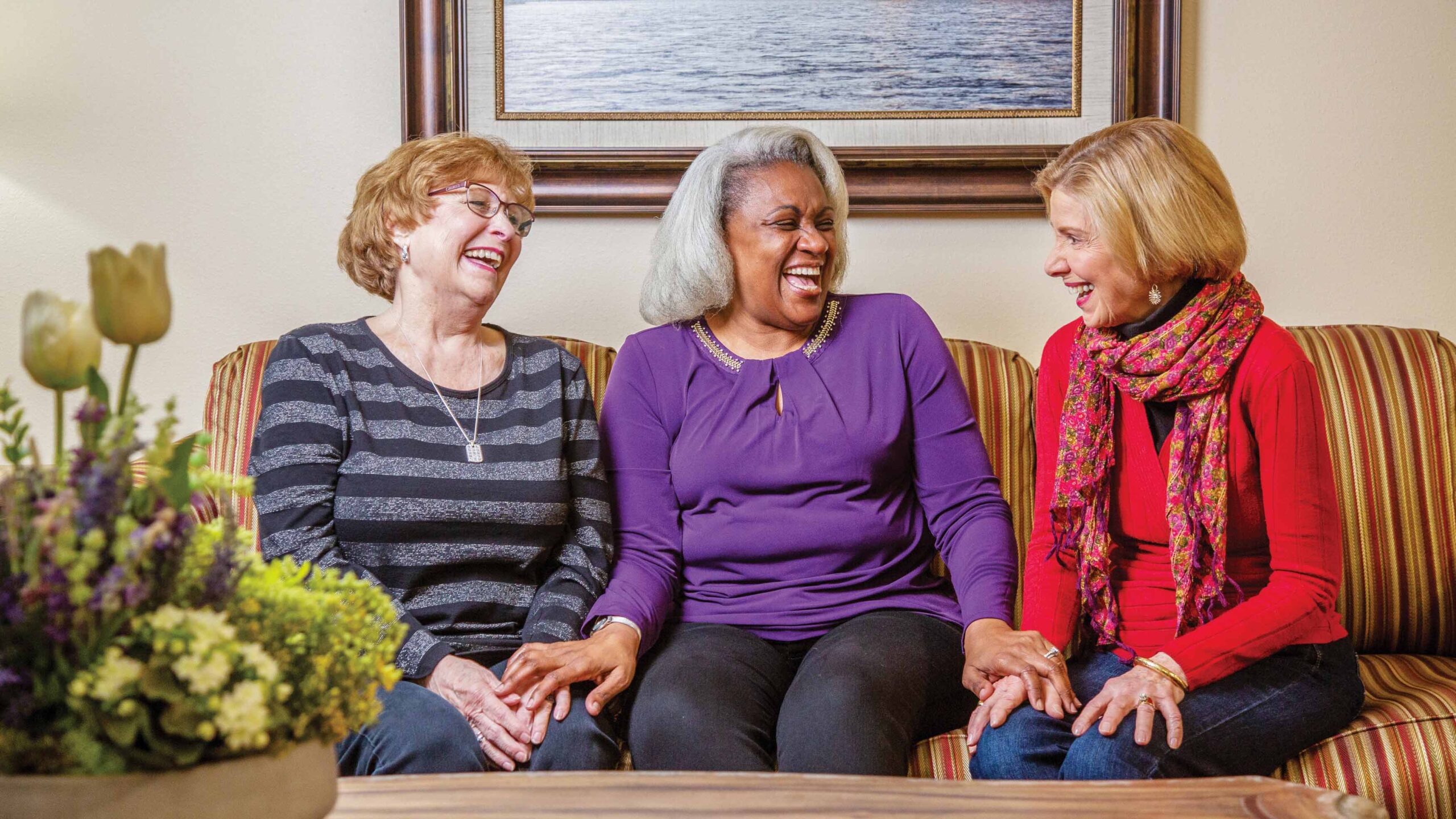 Three older women sit on a striped couch, smiling and laughing together. They are holding hands and appear to be enjoying a pleasant conversation. A flower arrangement is visible in the foreground.