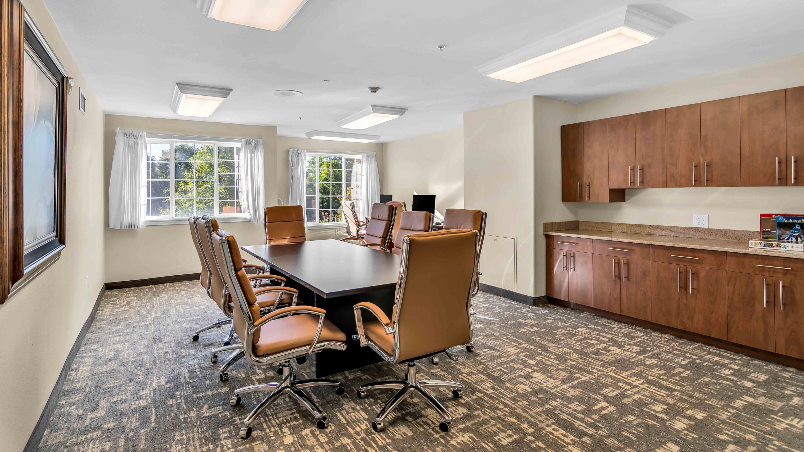 A modern conference room with a rectangular table surrounded by eight tan leather chairs, large windows letting in natural light, built-in wooden cabinets, and carpeted flooring.