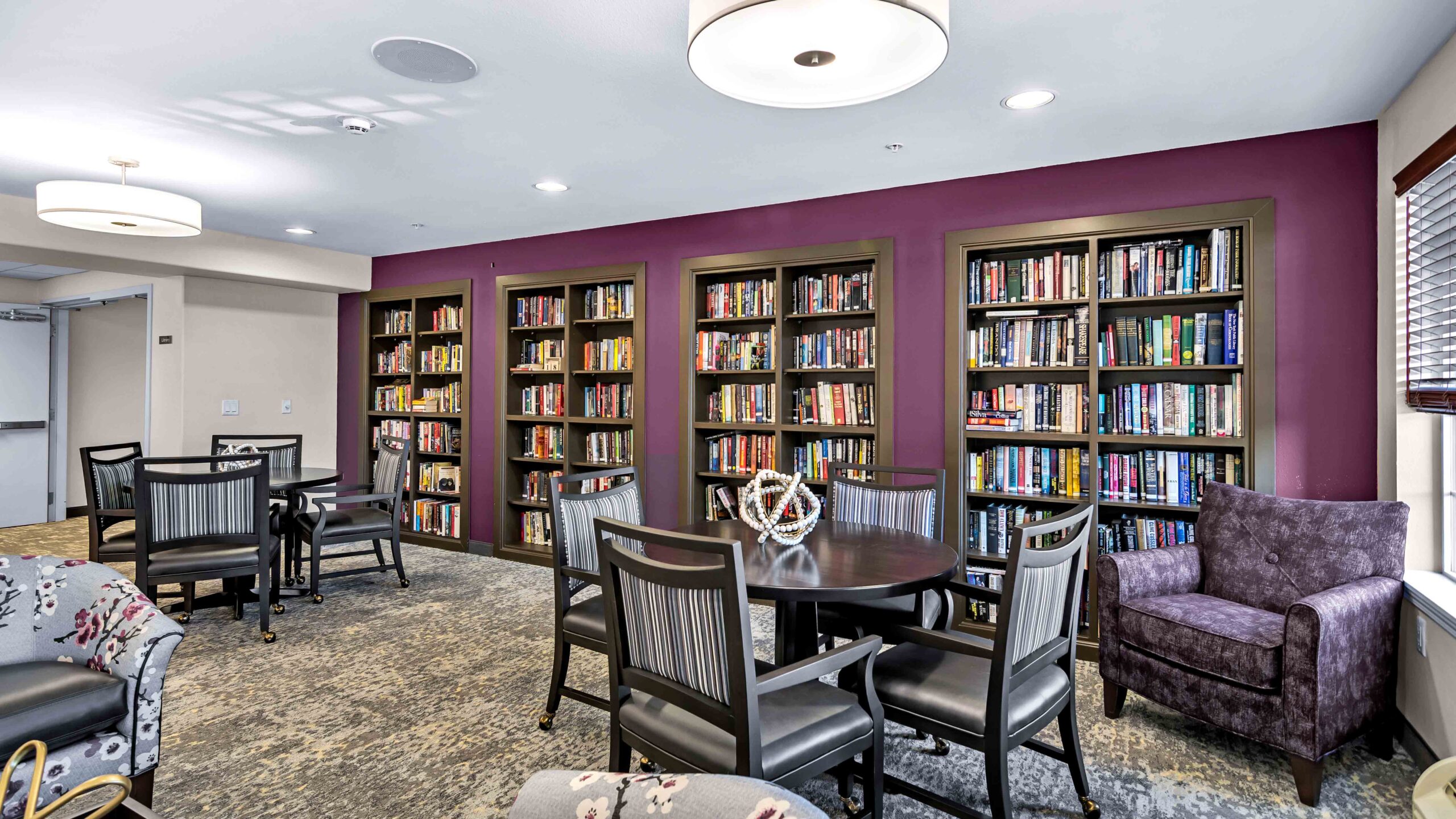 A cozy library room with bookshelves filled with books, round tables with chairs, a purple armchair, and modern lighting. The walls are accented with a deep purple color. Large windows let in natural light.