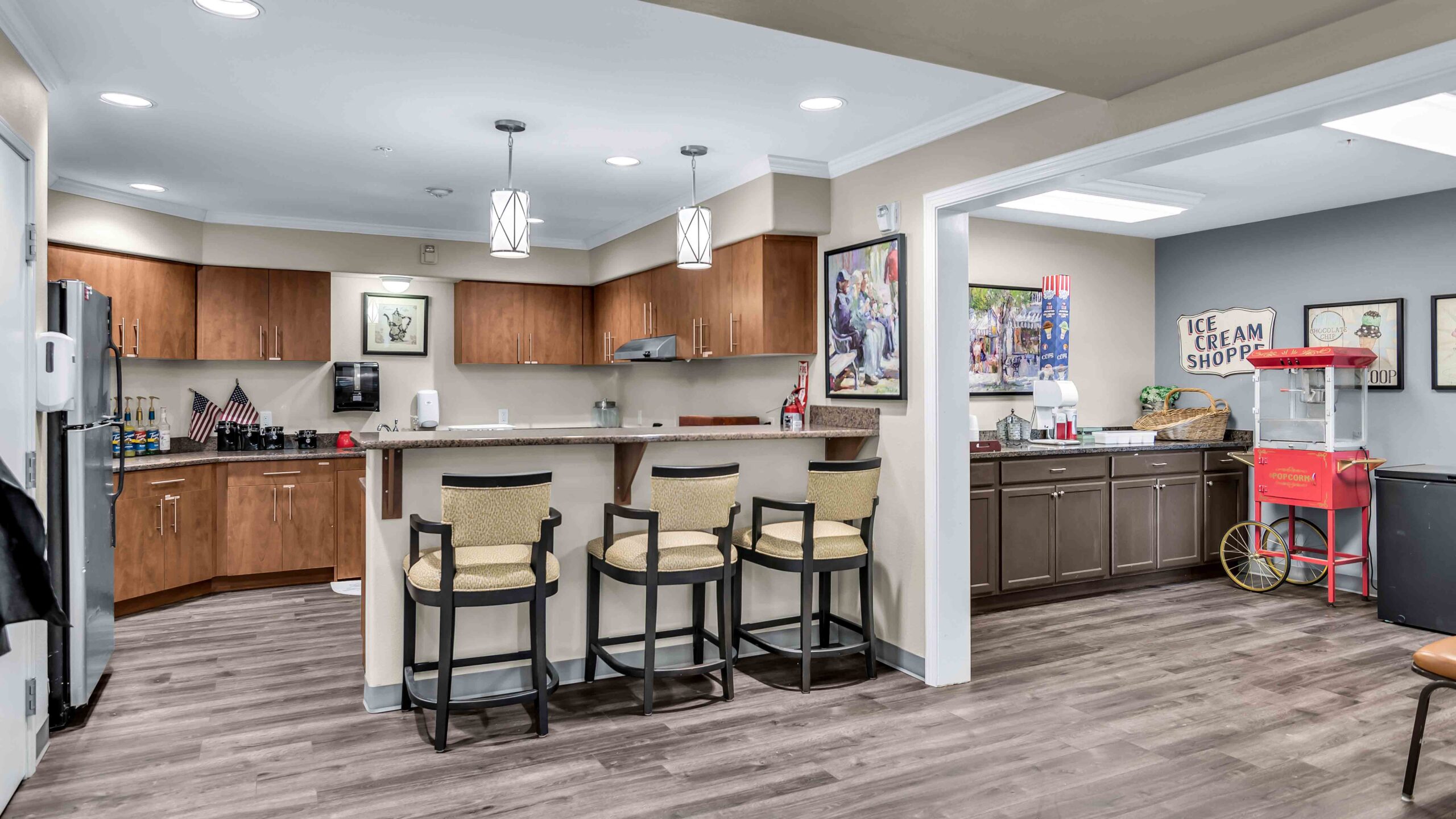 Modern kitchen with a breakfast bar and three chairs, wood cabinets, stainless steel appliances, and a section with an ice cream shop sign and a vintage popcorn cart on the right.