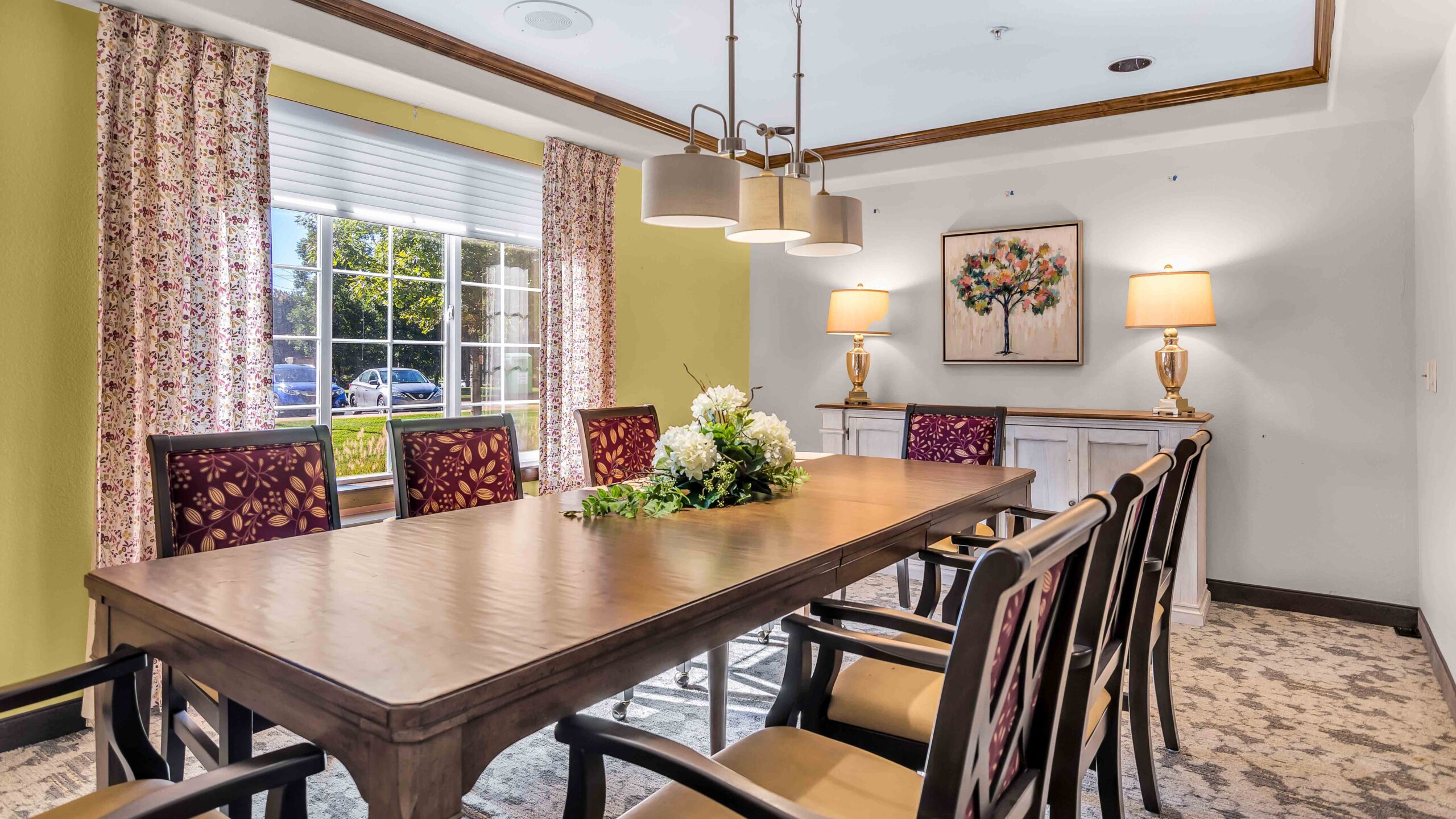 A dining room with a wooden table, eight chairs, floral centerpiece, large window with patterned curtains, two table lamps, a floral painting on the wall, and overhead lighting.