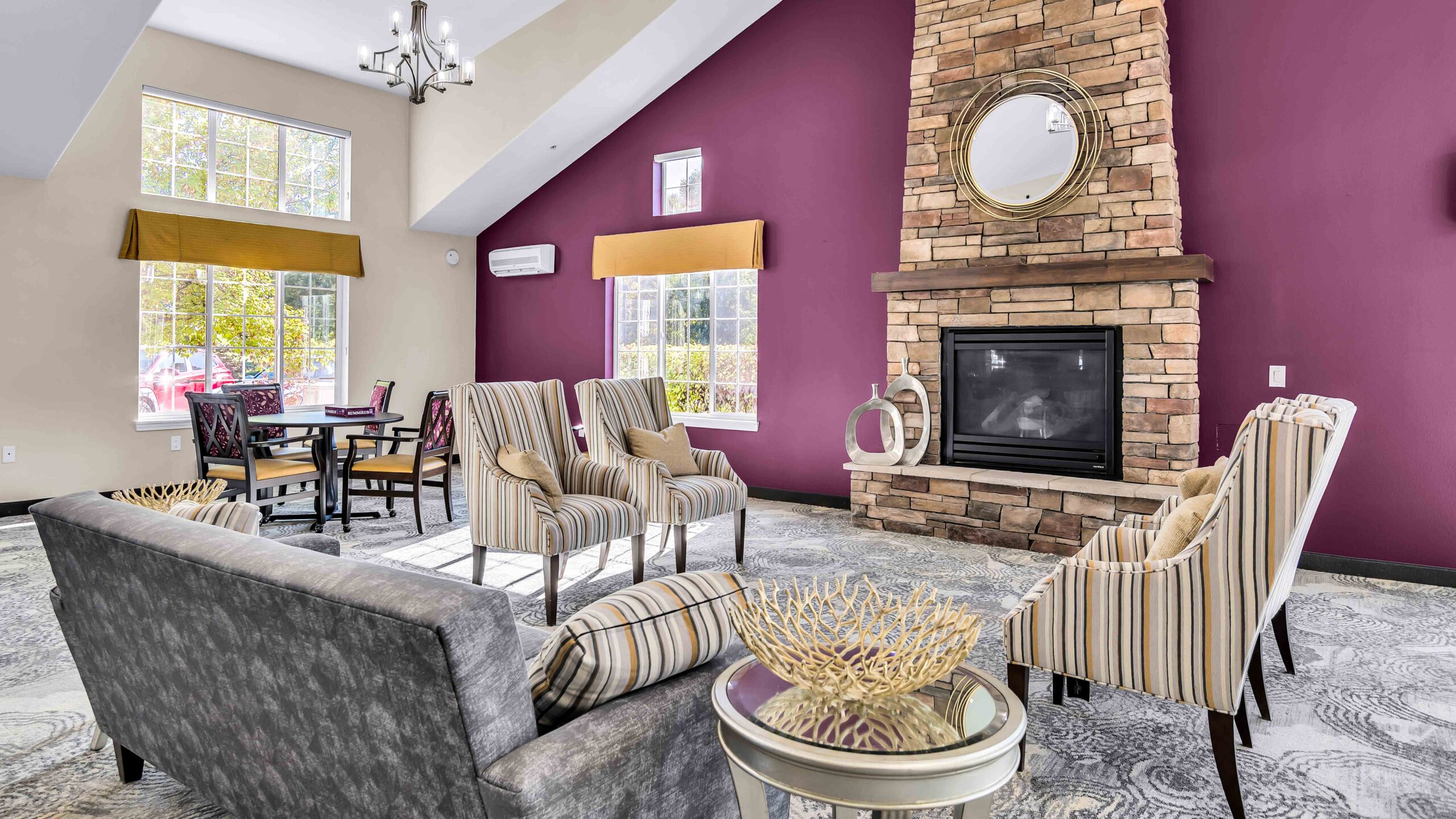 A modern living room with a stone fireplace, round mirror above it, striped armchairs, gray sofa, and a table with a decorative bowl. Large windows let in natural light, and the walls are painted purple and beige.